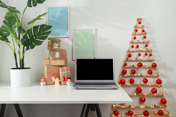 Christmas online shopping concept. Laptop and gifts on a white desk, original Christmas tree from branches in a home office. Morning sunshine.