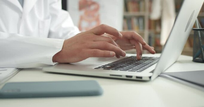 Crop view of man in white professional coat typing and filling in medical documentation. Male doctor working with laptop in office while sitting at table. Concept of routine and work