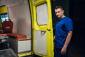 Corpsman in a blue uniform stands at the back of an ambulance car and looks at the camera.