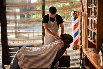Barber in apron trimming client beard in barbershop