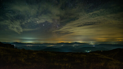 Night panorama of the mountainous area. Small villages glow in the night. Low clouds reflect light. The stars sparkle between the clouds