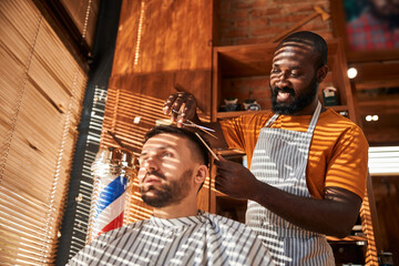 Smiling barber cutting client hair with scissors in barbershop