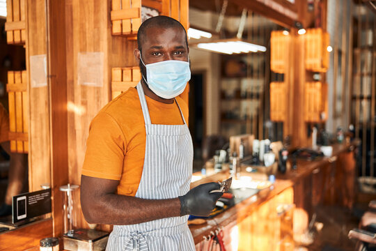Handsome male barber in medical mask standing in barbershop - Powered by Adobe