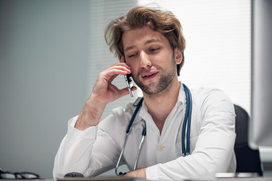 Male doctor talking over the cell phone, looking at his clipboard on the desk.