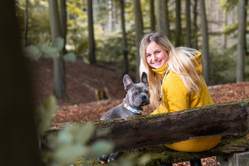 Happy, Smiling young blonde woman in yellow coat and her dog sitting on a bench in the autumn woods