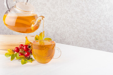 Herbal rose hips tea in teapot pouring into cup, autumn rosehip leaves and berry on white wood table with copy space.