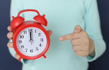 The Caucasian girl holds a red alarm clock in one hand , showing the time exactly twelve o'clock, and points the finger of the other hand in the direction of the clock. Specifying the time