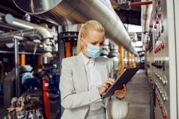 Dedicated blond female manager in suit with face mask standing in heating plant next to dashboard and using tablet to check on adjustments during corona virus outbreak.