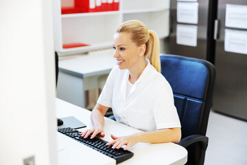 Smiling positive hardworking blond female lab assistant sitting in laboratory and entering data in computer.