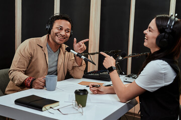 Portait of two cheerful radio hosts, man and woman smiling, pointing at each other, while moderating a live show in studio