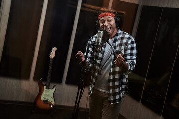 Portrait of young man, male artist looking focused, singing into a condenser microphone while recording a song in a professional studio