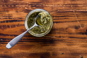 Top view photo of a jar of pesto genovese with a spoon, on a wooden rustic background.