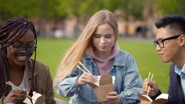Close up of diverse young man and women sitting on grass eating wok and chatting