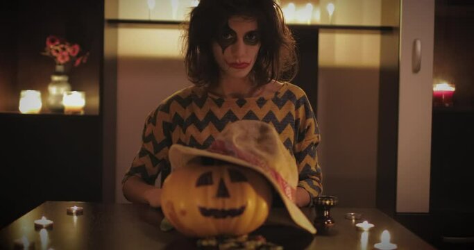 Young female with scary make up mask and knife sitting on the table with halloween pumpkin in front of her. Halloween day concept. 