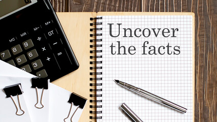 Notepad with text Uncover The Facts on the wooden background with clips, pen and calculator