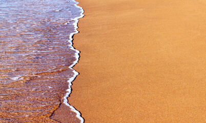 Close-up of the coast, clear sea blue water with shallow waves and a narrow strip of white foam washes the shore with a smooth yellow brown sand