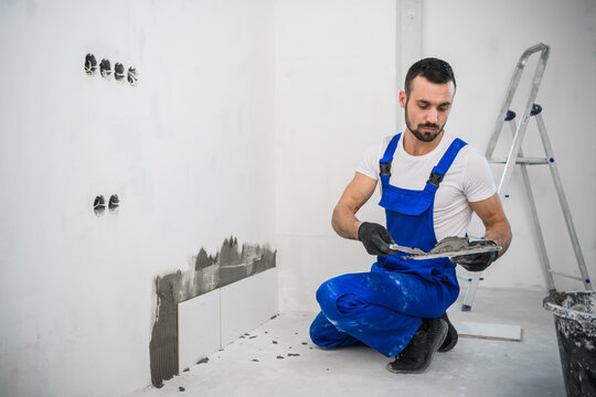The craftsman attaches the tiles to the wall with cement. He is wearing a blue uniform and black gloves