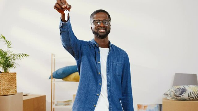 Close up of handsome african american man in denim shirt standing in new apartment. Man raises his hand up showing bunch of keys from new home.