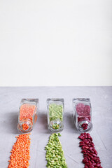 Dried green peas, orange lentils, red beans flowing out of crystal cleasr bottles on to grey concrete surface. Vertical. Copy space. Selective focus.
