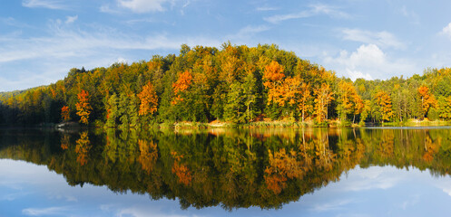Big panorama of autumnal colored trees by the lake, reflection of blue sky in water