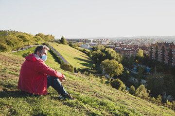 Fototapeta na wymiar A man in a red jacket and a medical mask sits on a green hill and looks at the city skyline. Traveling during a pandemic.