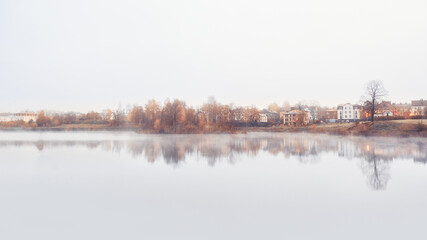 Panorama of a misty morning minimalistic landscape with a line of cottages and trees by the water in autumn.