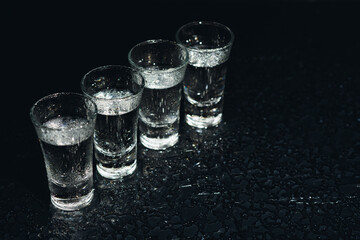 Vodka in shot glasses on black background, iced strong drink in misted glass