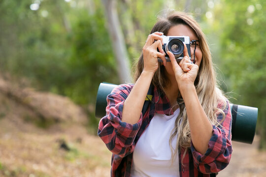Happy blonde woman taking photo of nature with camera and smiling. Caucasian long-haired traveler walking or hiking in forest. Blurred background. Tourism, adventure and summer vacation concept