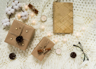 Fototapeta na wymiar Winter composition with an old book, glasses, gifts, pine cones, cinnamon, a cotton branch and glowing garlands on a light knitted blanket. Flat lay, top view. Warm cozy Christmas.