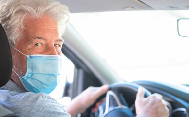 Portrait of a smiling white-haired senior man inside his car wearing surgical mask and looking back at camera, with hands of wheel