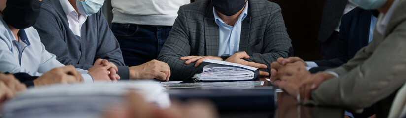 Officials or businessmen sit at a table next to a plethora of paper documents at a business meeting during a pandemic. Men in suits and medical masks. Reportage. Panorama.