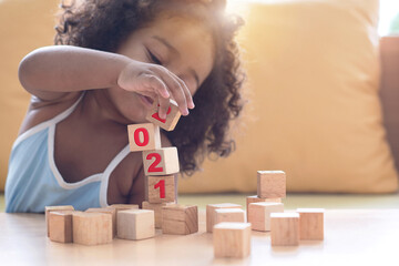 Fototapeta na wymiar Cute dark-skinned girl kid playing stacking wooden block with the year number 2021, in living room