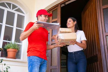 Cheerful Latin female client receiving parcels and smiling. Happy deliveryman delivering express order, talking with woman and holding clipboard. Express delivery service and post concept