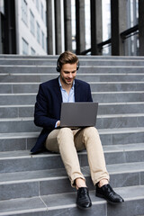Portrait of handsome smiling man in casual wear sitting on bench using laptop for online meeting, video call, video conference. Communication online with colleagues. Online education, remote working.