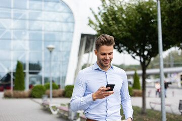 Handsome office worker holding smartphone and smiling. Happy young man using mobile phone apps, texting message, browsing internet, looking at smartphone. Coffee break near the business center.