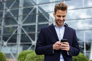 Handsome office worker holding smartphone and smiling. Happy young man using mobile phone apps, texting message, browsing internet, looking at smartphone. Coffee break near the business center.