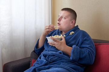 Fat man in a robe eating popcorn while sitting on the couch.