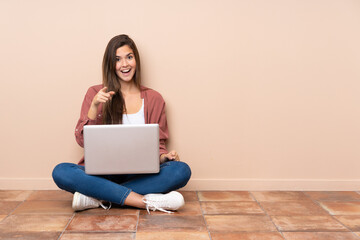 Teenager student girl sitting on the floor with a laptop surprised and pointing front