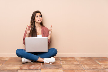 Teenager student girl sitting on the floor with a laptop with fingers crossing and wishing the best