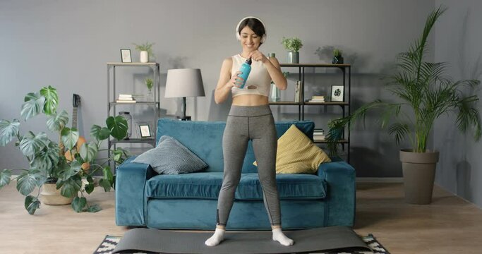 Cheerful Caucasian young woman in headphones drinking water while standing on yoga mat in living room. Working out at home. Sport and listening to music. Pretty happy female sipping dring as thirsty.