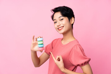 Young Asian girl over isolated pink background holding colorful French macarons and pointing it