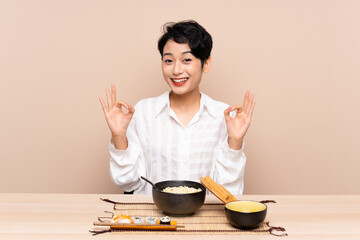 Young Asian girl in a table with bowl of noodles and sushi showing an ok sign with fingers