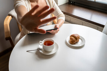 Woman showing stop gesture sign by hand having breakfast with cup of coffee working using tablet sitting near window in cafe at morning. Concept, coffee break