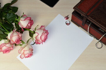 Empty greeting card on a light surface background. Next to it is a bouquet of roses, a jewelry box with a chain visible from it, a ring, a pendant with a pink stone in the form of a heart, a mobile ph