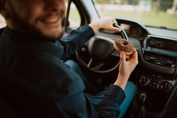 A close - up of hand of a young caucasian businessman holding a delicious chocolate doughnut and driving a car on the way to work. A sweet meal in transport