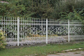 part of a white metal fence made of iron rods with a forged pattern in green grass outside