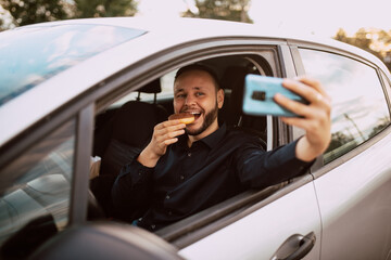 Naklejka premium A smiling happy caucasian man who takes a selfie while eating a donut with chocolate in the car. A sweet meal in the car
