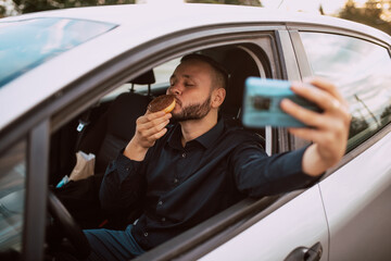 A young caucasian man who enjoys eating a donut with chocolate in the car and takes a selfie. A meal in the car