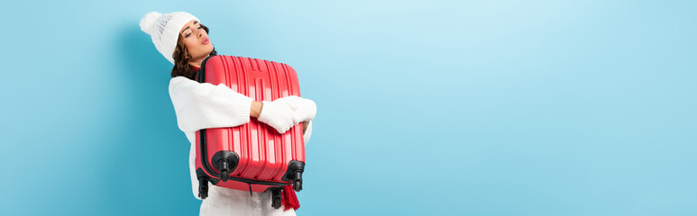 panoramic shot of young woman in winter outfit carrying heavy suitcase on blue
