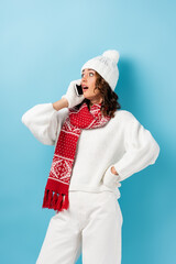 shocked young woman in white winter outfit talking on smartphone on blue